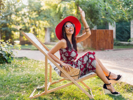 Elegante mujer hermosa sentada en la tumbona con traje de estilo tropical, agitando la mano, tendencia de la moda de verano, bolso de paja, sombrero rojo, gafas de sol, accesorios, sonriente, feliz humor, vacaciones